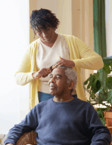 A young woman with dark skin, wearing a pale yellow shirt and cardigan stands while brushing the hair of an older man with dark skin in a dark blue sweater, who is sitting down.