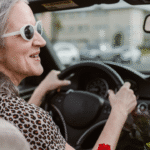 An older woman with light skin and grey hair wearing a leopard print shirt and white sunglasses drives a car.