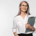 A light-skinned woman with shoulder-length hair is wearing a white button down shirt stands holding a clipboard in front of a white background