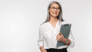 A light-skinned woman with shoulder-length hair is wearing a white button down shirt stands holding a clipboard in front of a white background