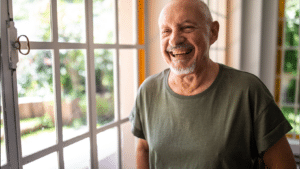 older man smiling while standing next to a window in a home
